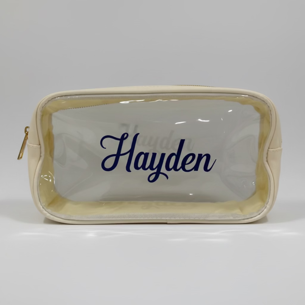 A clear pvc plastic cosmetic pouch with a cream-colored nylon border and a navy blue script name 'Hayden' printed on the front in permanent vinyl. zippered is gold 