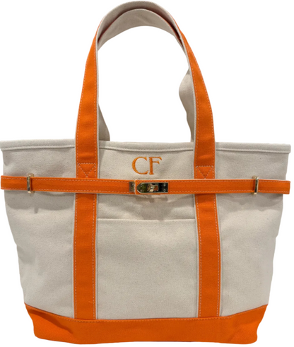 A canvas tote bag with orange trim, handles, and base, featuring a front pocket with an embroidered 'CF' monogram and a decorative orange strap across the middle with a gold buckle clasp.