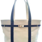 A natural color canvas tote bag with gold hardware and navy blue trim, double shoulder straps, and a gold-tone turn-lock closure. Outer pocket and inside ziper pocket and key chain hook.