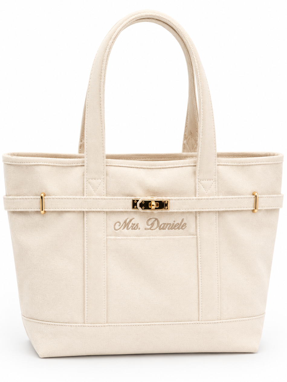Natural canvas tote bag with double top handles, a decorative horizontal strap with gold-tone hardware, and the name 'Mrs. Daniele' embroidered on the front pocket. Perfect engagement gift for the future bride. 